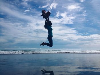 Woman holding bag while jumping on shore against sky