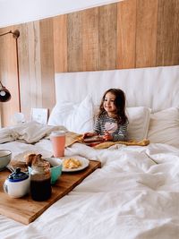 Full length of little girl sitting on bed having breakfast 