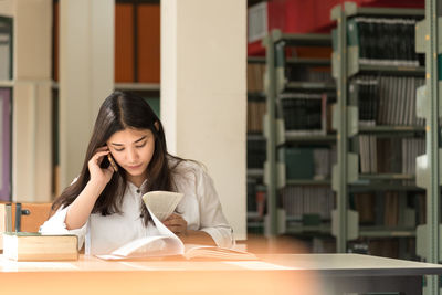 Young woman using phone while reading book on desk in library