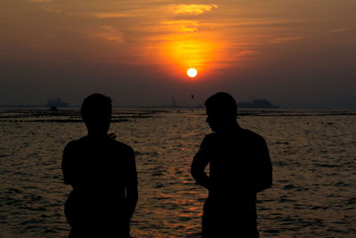 Rear view of silhouette friends standing at beach during sunset