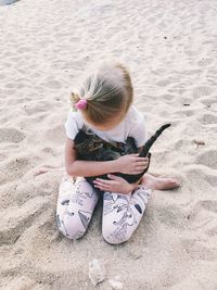 High angle view of girl carrying cat kneeling on sand at beach