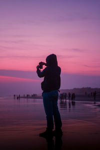 Silhouette man photographing by lake against sky during sunset