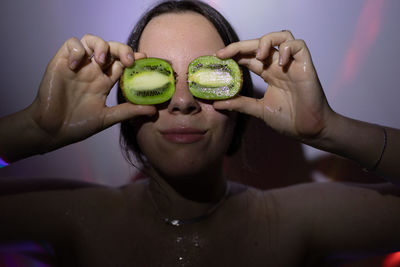 Young woman holding kiwi fruit against wall