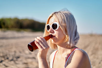 Young blond female in sunglasses sipping cold beer from bottle while spending sunny summer day on sandy beach
