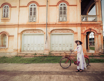 Portrait of young woman with bicycle standing on road against building