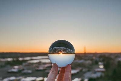 Close-up of hand holding crystal ball against sky during sunset