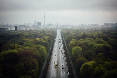 City street amidst trees leading towards fernsehturm in city against cloudy sky