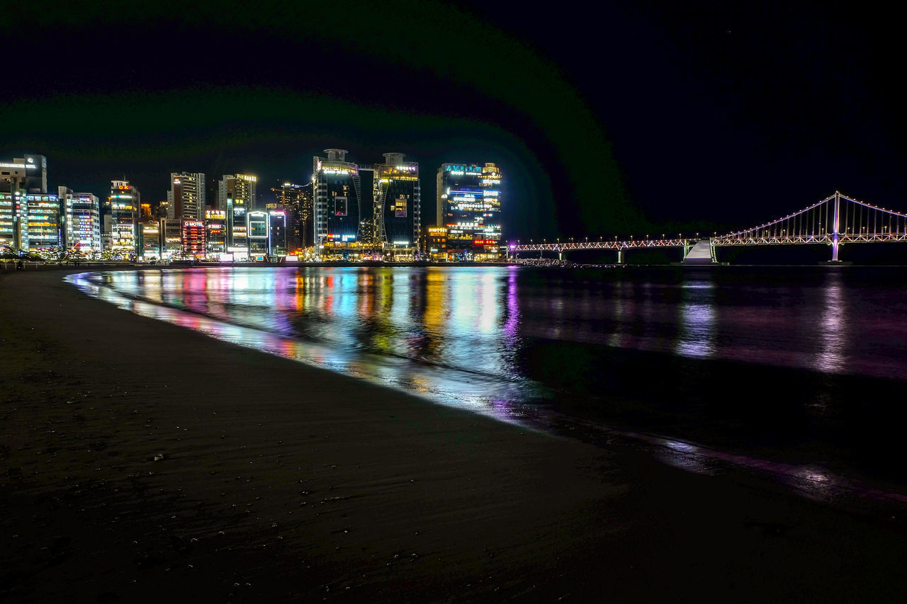 ILLUMINATED BRIDGE OVER RIVER BY BUILDINGS AGAINST SKY