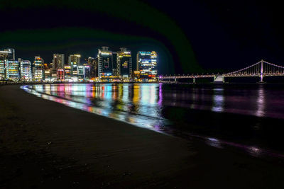 Illuminated bridge over river by buildings against sky at night