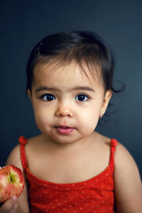 Baby girl in red tank top eating ripe red apple on black background in studio