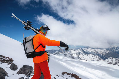 A young tall professional skier holds his skis while standing high in the mountains 