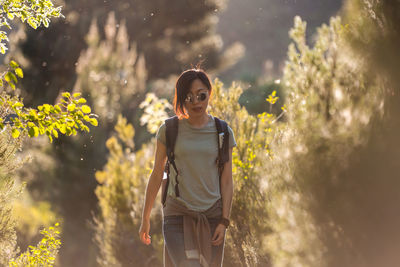 Asian female hiker in casual wear and sunglasses with rucksack strolling in nature with green plants during trekking on sunny summer day in valverde de los arroyos