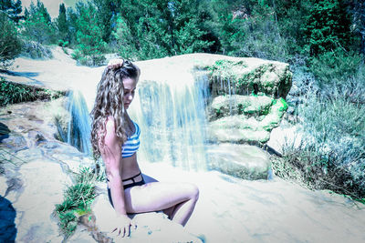 Side view of young woman sitting by waterfall in forest