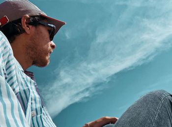 Man wearing sunglasses and cap while sitting at against blue sky