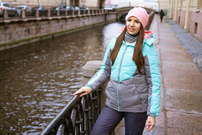 Young brunette woman in warm clothes and pink hat stands by river and smiles.