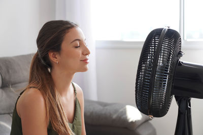 Relaxed happy young woman cooling down by ventilator at home on summer heat