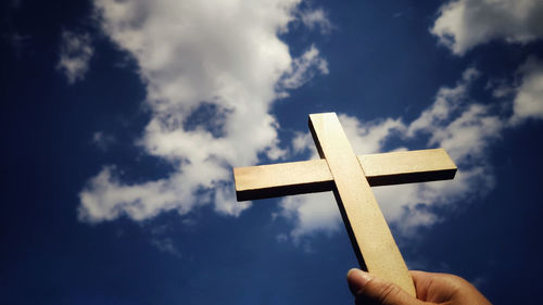 Low angle view of hand holding cross against sky