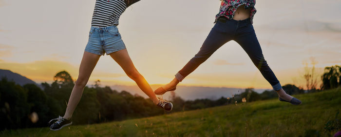 Low section of women jumping over field against sky during sunset