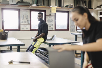 Smiling young male trainee leaning on table with coworker in foreground at workshop