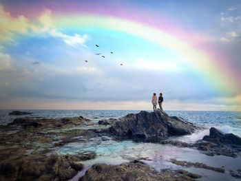 Man and woman standing on top of rock amidst sea looking at rainbow