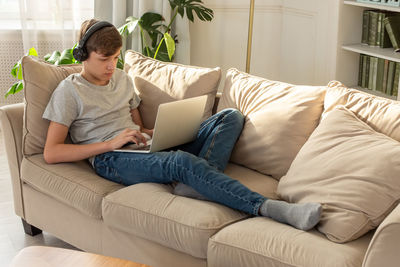 A teenager lying on a sofa in a room, wearing black headphones on his head, looks into a gray laptop