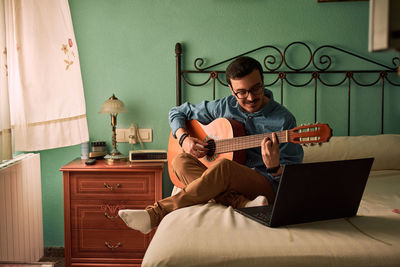 Young man practices with the guitar by looking at his laptop at home