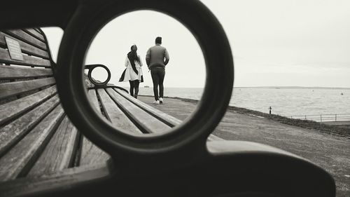 Rear view of couple walking on promenade seen through bench hole