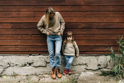 A young attractive mother and little daughter are standing near a wooden wall in the countryside.