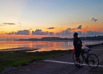 Rear view of man on bicycle by lake against sky during sunset