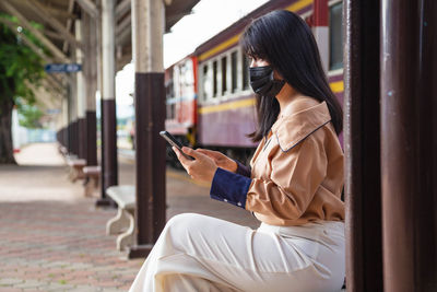 Side view of woman wearing flu mask using mobile phone at railroad station
