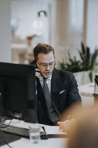 Young businessman talking on mobile phone while writing at computer desk in corporate office