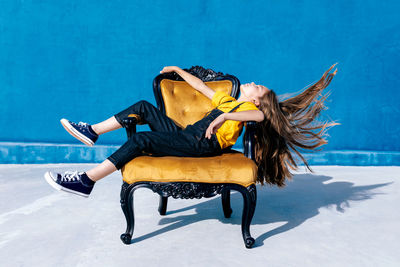 Side view of teen hipster sitting on chair and throwing hair on blue background