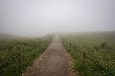 Dirt road amidst field against sky during foggy weather