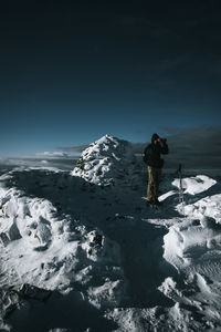 Young man looking through binoculars while standing on snow covered mountain against sky