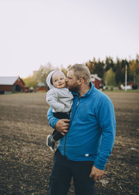 Father kissing toddler son standing at farm during sunset