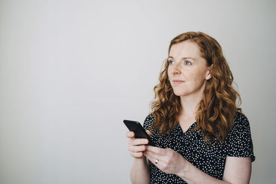 Thoughtful redhead businesswoman standing with smart phone against white wall at creative office