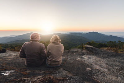 Rear view of people sitting on mountain against sky during sunset