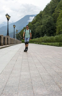 Young man in protective equipment roller skating along embankment against forest mountains in summer