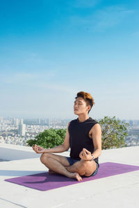 Young man sitting on seat in city against sky