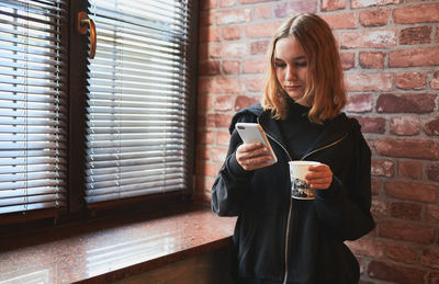 Woman texting on smartphone, drinking coffee while taking a break in office standing at window