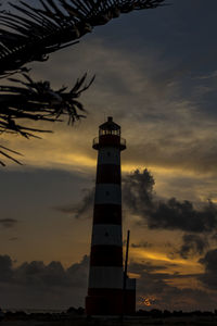 Silhouette lighthouse by building against sky during sunset