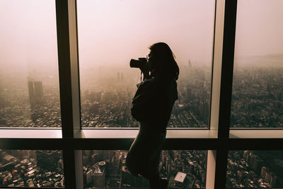 Side view woman photographing through window against cityscape