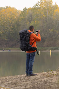 Portrait of traveler man at lake in autumn taking a photo with the digital camera and orange jacket