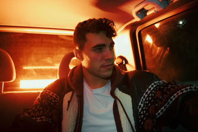 Side view of confident young male millennial sitting in modern car with red neon illumination at night and looking away