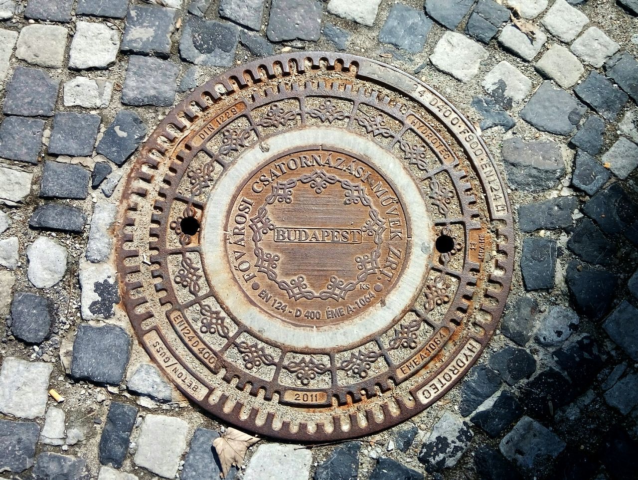 circle, geometric shape, shape, street, manhole, text, no people, day, metal, craft, high angle view, design, western script, pattern, stone, city, sewer, architecture, art and craft, close-up, outdoors, paving stone, carving