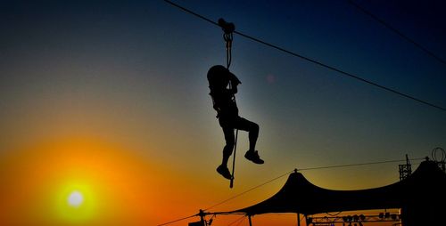 Low angle view of silhouette man zip lining against clear sky during sunset