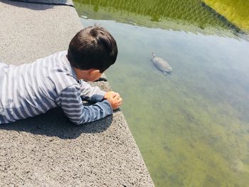 High angle view of boy looking at turtle swimming in lake while lying on retaining wall