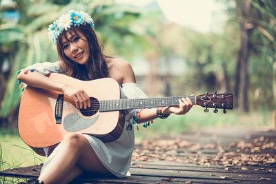 Portrait of smiling woman playing guitar while sitting on boardwalk at park