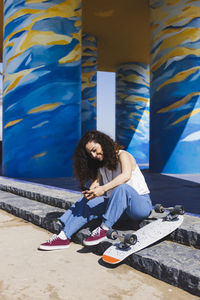 Full body side view of female in casual clothes sitting on stairs near skateboard browsing on smartphone on sunny street during training