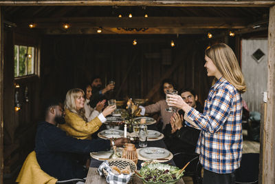 Smiling woman toasting drink with male and female friends while standing by dining table party